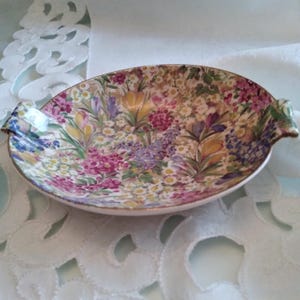 May include: A shallow, oval-shaped ceramic dish featuring a vibrant, multi-colored floral pattern with yellow, purple, pink, and white blossoms. The dish has small, integrated handles on each side and a thin gold-toned rim along the edge.