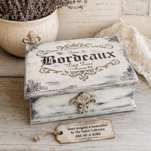 May include: A rectangular wooden box with a distressed white and grey finish, featuring a black printed label that reads Vins de Bordeaux Petit Paris 14 France 53. The box has a decorative metal fleur-de-lis latch and a hanging paper tag.