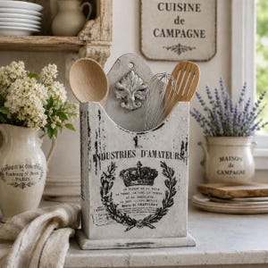 May include: A white distressed wooden utensil holder featuring a black printed graphic with a crown, laurel wreath, and the text "DES INDUSTRIES D'AMATEURS". It contains a wooden spoon, a wooden spatula, and a metal whisk, with a decorative crest.
