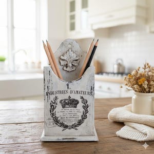 May include: A distressed white pencil holder with a decorative floral design and text. The holder contains several pencils. The holder is made of wood and sits on a wooden surface. The overall theme is vintage and rustic.