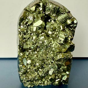 May include: A metallic gold pyrite mineral specimen featuring a cluster of naturally formed cubic crystals. The stone has a rounded top edge and a dense, textured surface with reflective facets that catch the light, displaying a brassy yellow hue.