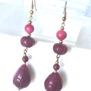 May include: A pair of dangle earrings with three pink and purple beads on each earring. The beads are carved with a floral pattern and are connected by gold-toned metal.