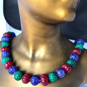 May include: A colorful necklace featuring large, ribbed beads in shades of red, blue, and green. The beads are strung together to create a statement piece. The necklace is displayed on a gold-toned mannequin.