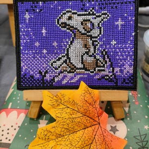 Könnte beinhalten: Eine violette und weiße Kreuzstich-Stickerei mit einer Comicfigur vor einem sternenübersäten Hintergrund. Die Stickerei ist gerahmt und auf einem kleinen Holzständer ausgestellt.