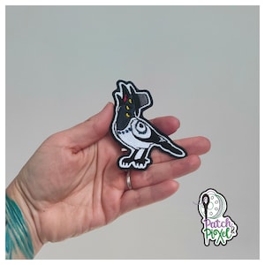 Può includere: Toppa ricamata raffigurante un uccello stilizzato in bianco e nero con dettagli gialli e rossi. La toppa, con bordo nero, è tenuta in mano. Il disegno dell'uccello ha un caratteristico motivo oculare. Per abbigliamento o accessori.