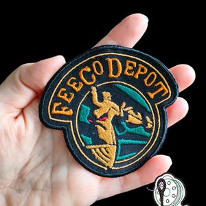 Op de afbeelding: Geborduurde patch met de tekst "EECO DEPOT" in gouden letters op een zwarte achtergrond. De patch heeft een gestileerde gouden figuur, met groene en rode accenten, en een cirkelvormig ontwerp. De patch wordt in een hand gehouden.
