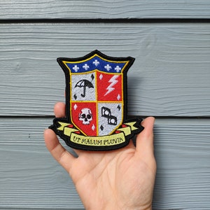 Könnte beinhalten: Ein gestickter Patch in Form eines Schildes mit einem roten, weißen und schwarzen Wappen. Das Wappen zeigt einen Regenschirm, einen Blitz, einen Totenkopf und Würfel. Der Text "UT MALUM PLUVIA" ist auf einem gelben Banner unter dem Wappen geschrieben.