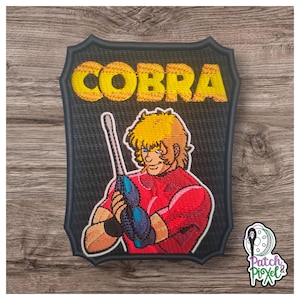 May include: Embroidered patch featuring the word "COBRA" in yellow block letters above an illustration of a character holding a weapon. The character is wearing a red shirt and has yellow hair. The patch has a black background.