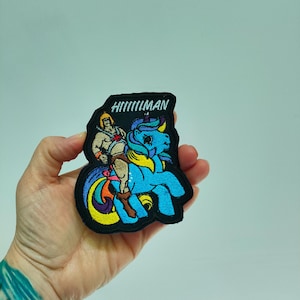 May include: An embroidered patch with a black border featuring a muscular figure in a gold helmet riding a bright blue unicorn with a rainbow-colored mane and tail. The text HIIIIIMAN is stitched in white at the top of the patch design.