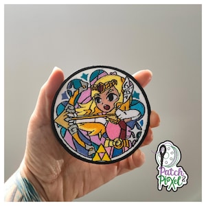 Könnte beinhalten: Runder, gestickter Aufnäher mit einer Zeichentrickfigur mit blonden Haaren, einer Krone und Pfeil und Bogen. Das Design umfasst rosa, blaue, gelbe und weiße Farben mit einem schwarzen Rand. Der Aufnäher hat den Text "Patch Pixel" unten rechts.