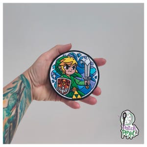 Könnte beinhalten: Runder, gestickter Aufnäher mit einer Cartoonfigur mit Schwert und Schild in Grün-, Blau- und Gelbtönen. Der Aufnäher hat einen schwarzen Rand und wird in einer Hand gehalten. Der Aufnäher hat einen Durchmesser von ca. 7,6 cm.
