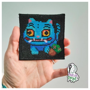 Peut inclure: Un écusson carré noir avec un monstre bleu brodé au point de croix, aux yeux orange et un petit objet vert et marron. L'écusson est tenu dans une main. Le texte "Patch Pixel&" est dans le coin inférieur droit.