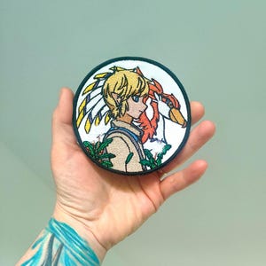 Peut inclure: Écusson brodé rond représentant un personnage aux cheveux blonds et un oiseau rouge. L'écusson a une bordure noire et un fond blanc. Le personnage porte une tenue beige avec des accents bleus.