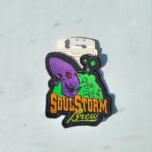Könnte beinhalten: Ein gestickter Aufnäher in Schwarz, Grün, Lila und Orange mit dem Text "Soulstorm Brew".