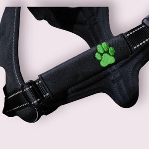May include: Black dog harness with a green paw print embroidered on the chest strap. The harness has a reflective strip and adjustable straps.