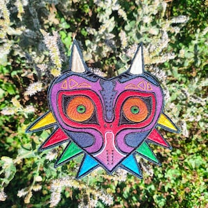 Könnte beinhalten: Gestickter Aufnäher mit dem Majora's Mask-Design aus der The Legend of Zelda-Reihe. Der Aufnäher hat eine Herzform mit rotem Gesicht, orangefarbenen Augen und farbenfrohen Akzenten. Der Aufnäher ist von grünem Laub umgeben.