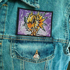 May include: A rectangular embroidered patch featuring a stylized yellow and orange lightning bolt design against a purple background. The patch is bordered by a black frame and is affixed to a blue denim jacket.