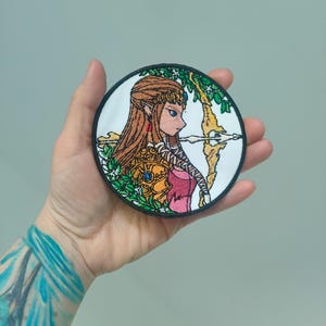 Peut inclure: Écusson brodé rond représentant un personnage aux longs cheveux bruns, aux yeux bleus et vêtu d'une tenue rose et dorée, tenant un arc et une flèche. L'écusson a une bordure noire et un fond blanc avec des feuilles vertes.