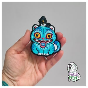 Könnte beinhalten: Gestickter Aufnäher mit einer blauen Katze mit großen gelben Augen, einem schwarzen Hut und einem breiten Grinsen. Der Aufnäher hat einen schwarzen Rand und wird in einer Hand gehalten. Das Katzendesign ist detailliert mit verschiedenen Blau- und Weißtönen.