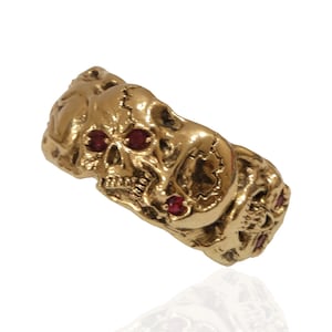 May include: A gold-colored ring featuring a detailed skull design. The skull's eye sockets are adorned with small, red gemstones. The ring has a textured surface and a gothic aesthetic.
