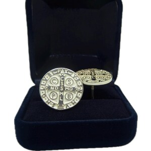 May include: A pair of silver cufflinks featuring a Saint Benedict medal design. The medals are round and have a black cross in the center with the words "C.S.S.M.L.  N.D.S.M.D.  P.A.X." around the edge. The cufflinks are in a black velvet box.