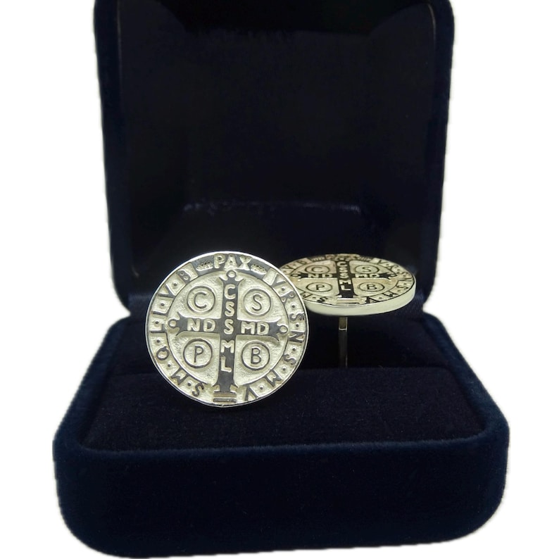 May include: A pair of silver cufflinks featuring a Saint Benedict medal design. The medals are round and have a black cross in the center with the words "C.S.S.M.L.  N.D.S.M.D.  P.A.X." around the edge. The cufflinks are in a black velvet box.