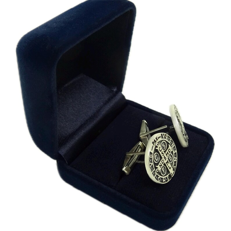 May include: A pair of silver cufflinks in a blue velvet box. The cufflinks feature a white enamel design with a religious symbol and inscription.