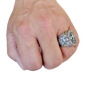 K&ouml;nnte beinhalten: Ein silberner Ring mit einem komplizierten Design, das ein Wappen mit einem Kreuz, einem Schild und einer Krone zeigt. Der Ring wird an einem Finger getragen.
