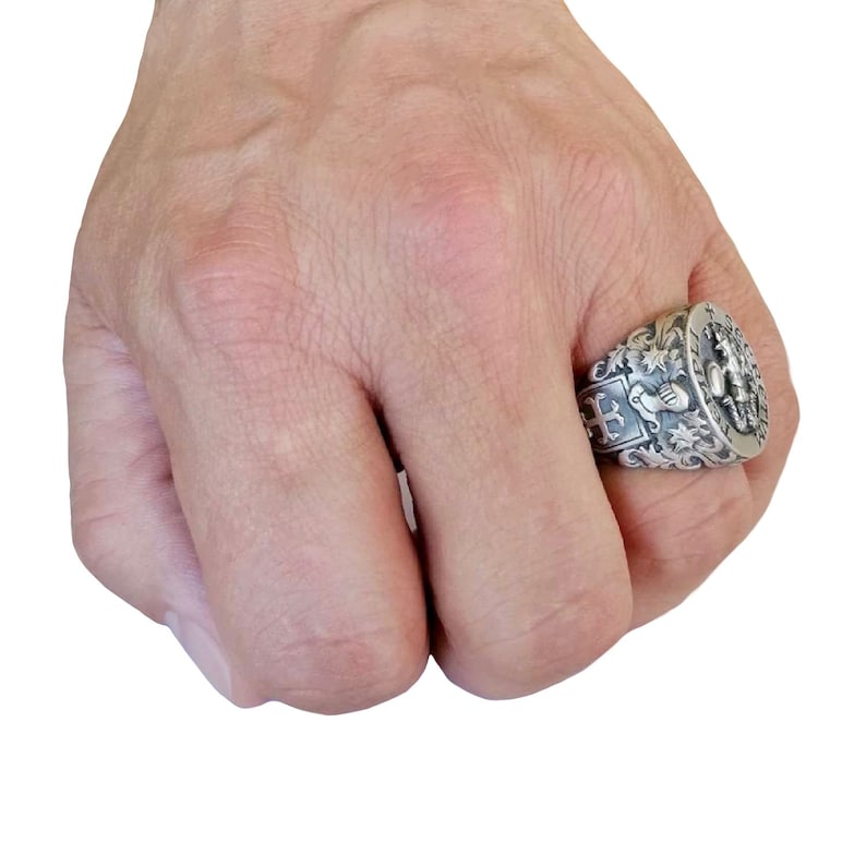 K&ouml;nnte beinhalten: Ein silberner Ring mit einem komplizierten Design, das ein Wappen mit einem Kreuz, einem Schild und einer Krone zeigt. Der Ring wird an einem Finger getragen.