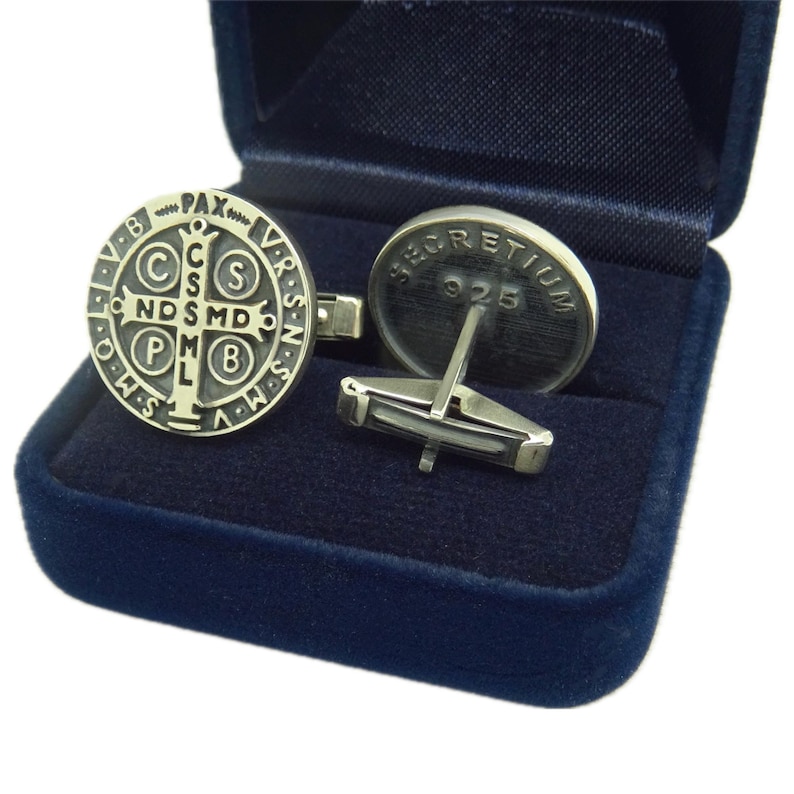 May include: A pair of silver cufflinks in a blue velvet box. One cufflink features a silver St. Christopher medal with the inscription "PAX VR・S・N・S・MV C ND SMD PHO SMQ." The other cufflink has the inscription "SECRETIVM 925".