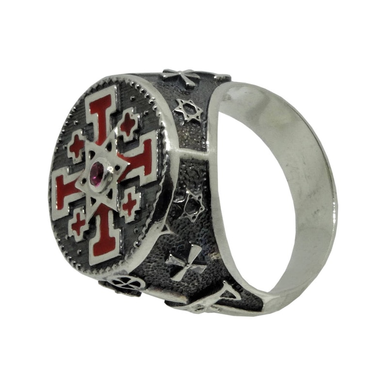 May include: A silver ring with a red and black enamel design featuring a cross and a star. The ring has a textured band with additional symbols.