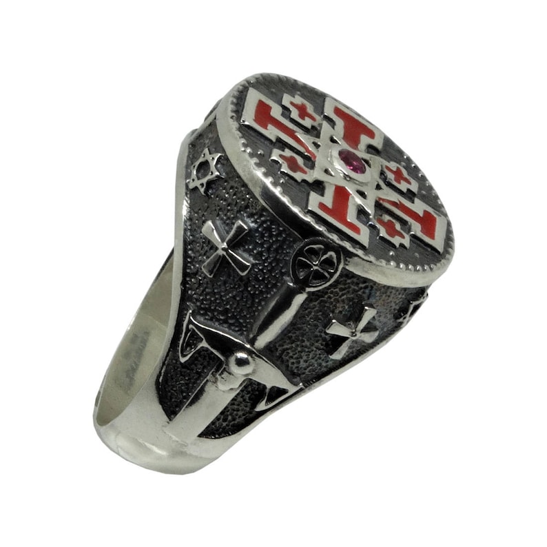May include: A silver ring with a red and black enamel design. The ring features a cross with a red stone in the center. The ring is detailed with other symbols.