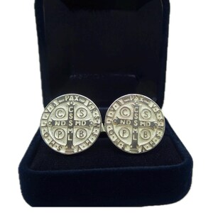 May include: A pair of silver cufflinks featuring a Saint Benedict medal design. The medals are round and have a raised cross in the center with the words "PAX" and "C.S.S.M.L." around the edge.
