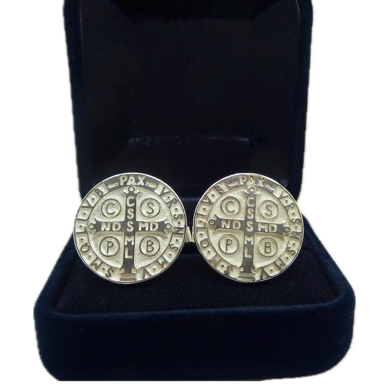 May include: A pair of silver cufflinks featuring a Saint Benedict medal design. The medals are round and have a raised cross in the center with the words "PAX" and "C.S.S.M.L." around the edge.