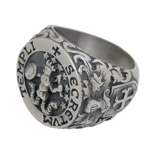 K&ouml;nnte beinhalten: Ein silberner Ring mit aufwendigen Gravuren. Der Ring zeigt ein Wappen mit einem Kreuz und einem Fabelwesen, umgeben von floralen Designs. Die Inschrift "TEMPLI SECRETVM" ist auf dem Ring zu sehen.