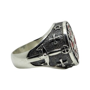 May include: A silver ring with a detailed design featuring a cross, a star of David, and other symbols. The ring has a textured surface and a red enamel inlay.