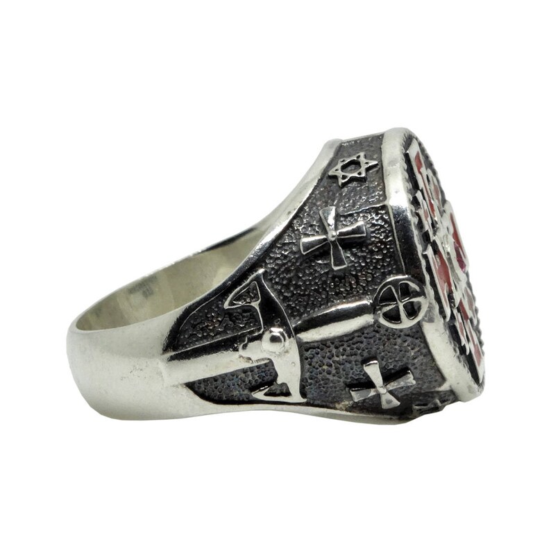May include: A silver ring with a detailed design featuring a cross, a star of David, and other symbols. The ring has a textured surface and a red enamel inlay.