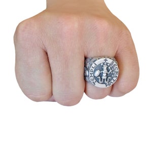 K&ouml;nnte beinhalten: Ein silberner Ring mit einem runden Design, das einen Ritter mit einem Schild und einem Schwert zeigt. Der Ring ist mit den Worten "TEMPLI SECRETVM" beschriftet.