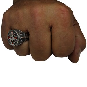 May include: A silver ring with a red and black enamel design featuring a cross with eight points. The ring is worn on a person's finger.