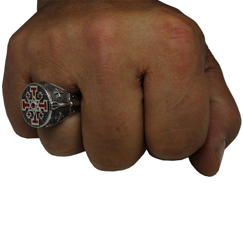 May include: A silver ring with a red and black enamel design featuring a cross with eight points. The ring is worn on a person's finger.