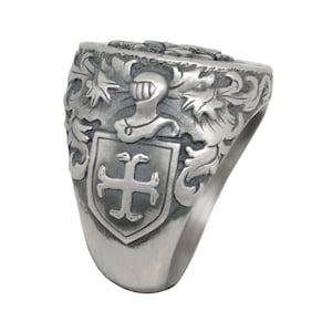 K&ouml;nnte beinhalten: Ein silberner Ring mit einem detaillierten Design, das einen Schild mit einem Kreuz, einen Helm und verzierte Ranken zeigt.