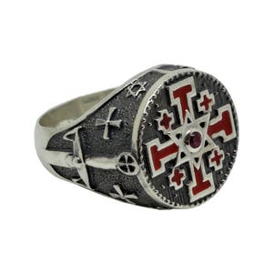 May include: A silver ring with a red enamel cross design. The cross is surrounded by smaller crosses and a star with a red gemstone in the center.