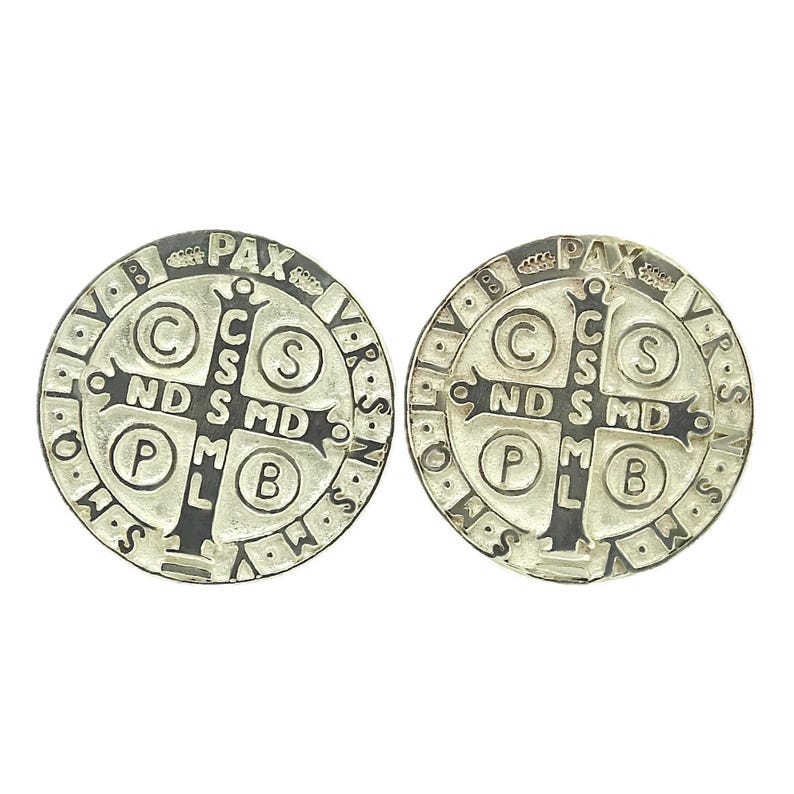 May include: Two silver-colored Saint Benedict medals with black lettering. The front of each medal features a cross with the letters 'C.S.S.M.L.' and 'N.D.S.M.D.' around the cross. The back of each medal features the words 'PAX' and 'VBS' around the edge.
