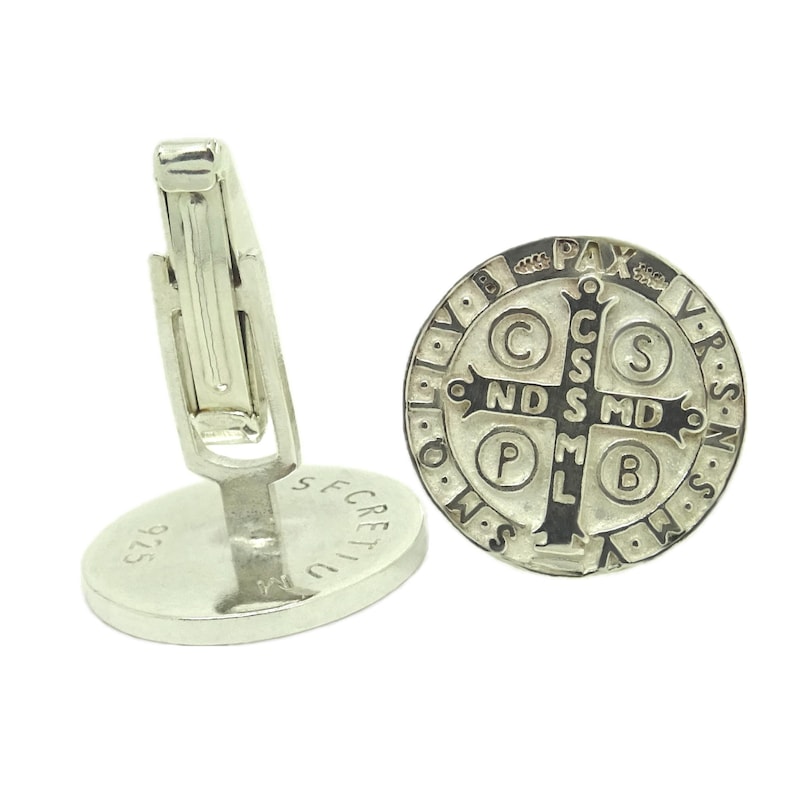 May include: Silver cufflinks featuring a Saint Benedict medal with a black cross and inscription. The back of the cufflinks are engraved with "925" and "SECRETIC".
