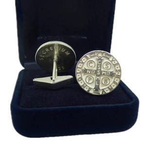 May include: A pair of silver cufflinks in a blue velvet box. One cufflink features a round design with the words "Secretum 25". The other cufflink features a round design with a cross and the words "In Pax" and "C.S.S.M.D.P.L.B."