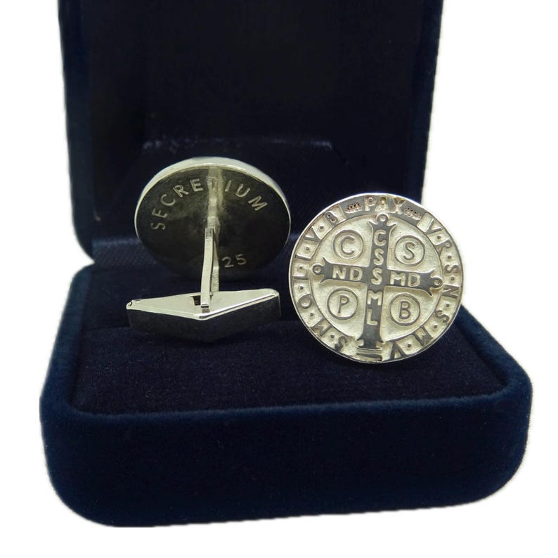 May include: A pair of silver cufflinks in a blue velvet box. One cufflink features a round design with the words "Secretum 25". The other cufflink features a round design with a cross and the words "In Pax" and "C.S.S.M.D.P.L.B."
