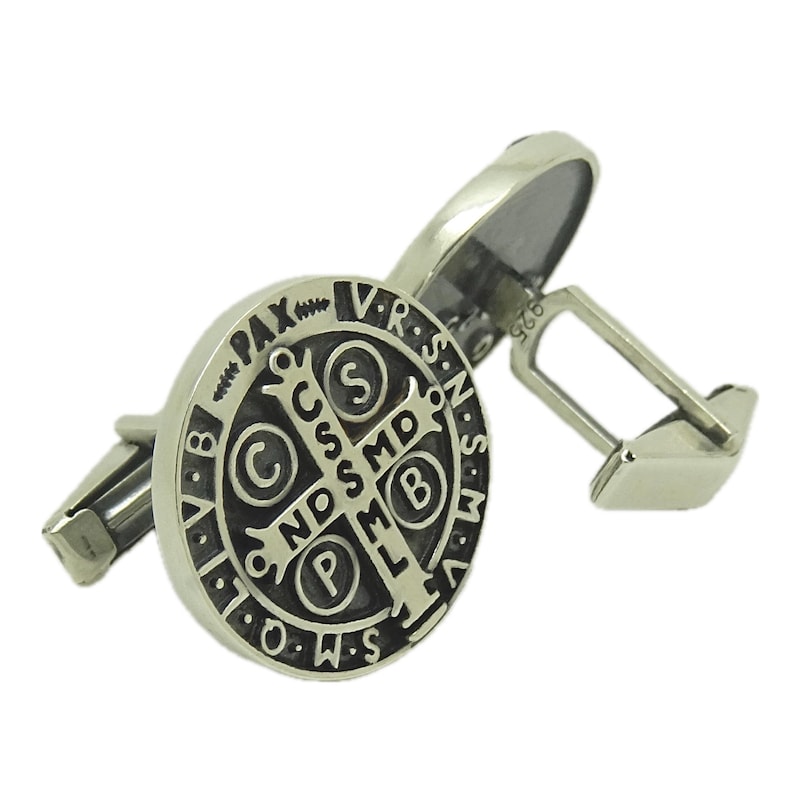 May include: A pair of silver cufflinks featuring a black and silver Saint Benedict medal. The medal has the inscription "V.R.S.N.S.M.D.O.W.S.P.L.M.B.C.C.S.S.M.D. PAX"