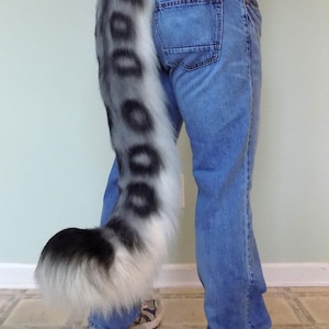 May include: A grey and white faux fur tail with large, dark grey spots. The tail is attached to the back of a person wearing blue jeans. The tail has a fluffy, rounded end.