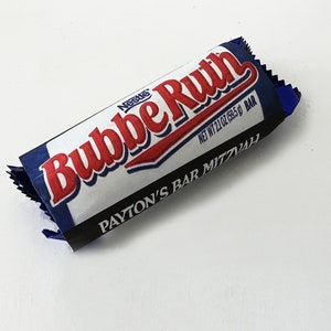 May include: A wrapped Nestle Bubbe Ruth candy bar. The wrapper is white with a blue border and the words "Bubbe Ruth" in red script. The bar is 2.1 oz (59.5 g) and says "Payton's Bar Mitzvah" on the wrapper.