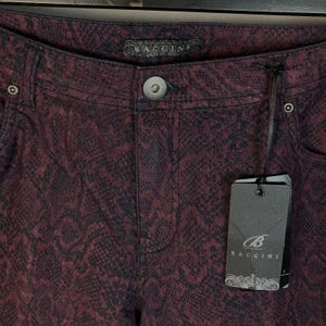 May include: Dark burgundy and black pants featuring a snakeskin pattern. The pants have a central button closure, metal rivets at the pockets, and a black brand label reading BACCINI. A black rectangular hang tag with the BACCINI logo is attached.